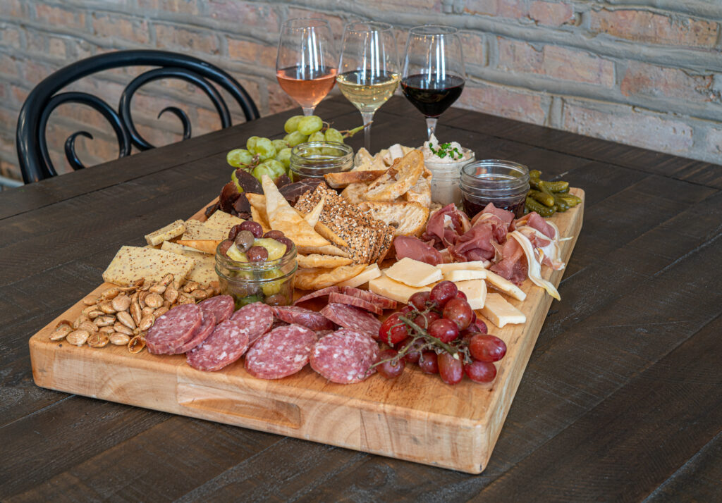Chacuterie board on a dark wood table, an assortment of cheeses, meats, snacks, and pairings of three wines.